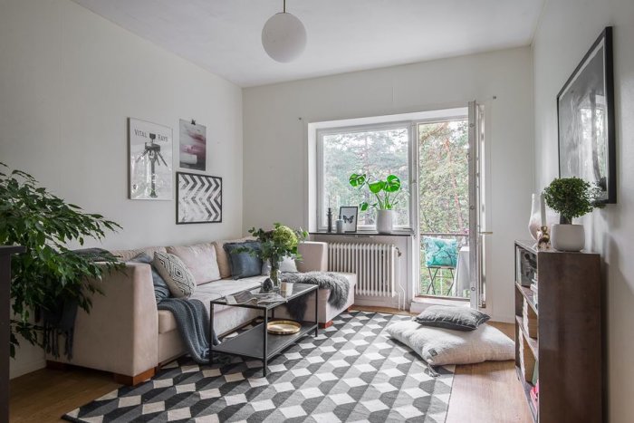 Vardagsrumsinteriör med beige soffa, soffbord, mönstrad matta, växter och fönster med utsikt mot grönska.