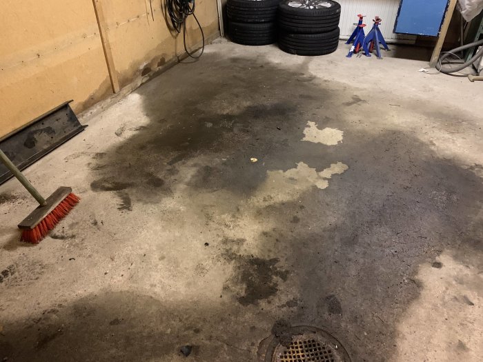 Betonggolv i ett garage med ojämnt fall och tydliga våta fläckar nära en av väggarna, en kvastskaft och däck syns i bakgrunden.