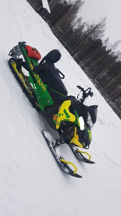 Grön och gul snöskoter parkerad på snötäckt mark mot skogsbakgrund.