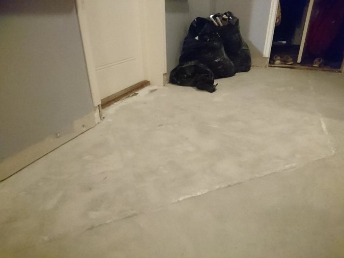 Slipad betonggolv i hall med synliga reglar, plastskydd vid dörröppning och svarta sopsäckar i hörnet.