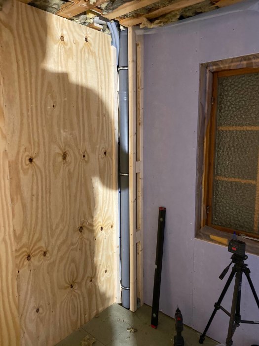 Renoveringsarbete visande plywoodvägg, avloppsrör, verktyg och en bygg-laser på ett stativ.