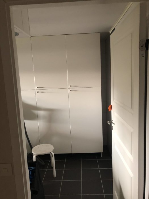 Frysskåp integrerat i vit skåpsinredning i en tvättstuga, med en tallriksventil på väggen och en vit pall på golvet.