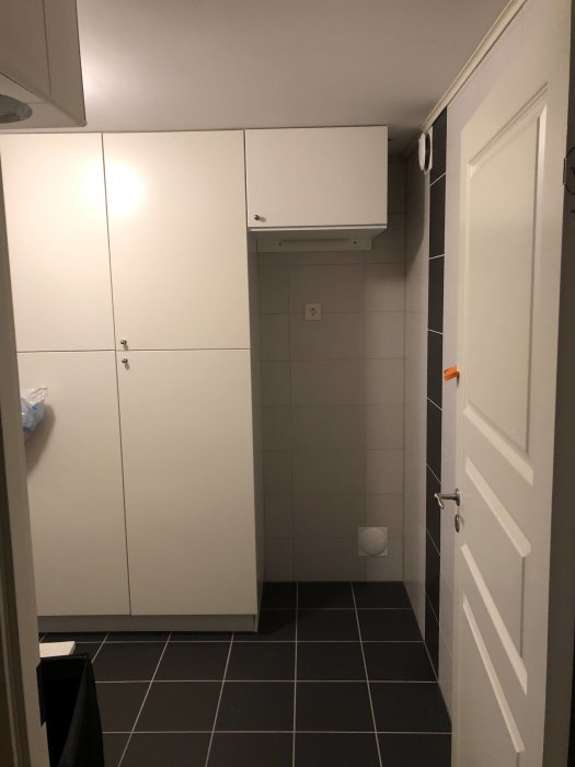 En tvättstuga med vit frys högt monterad, skåp och en dold tallriksventil på kakelväggen.
