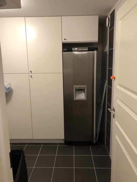 Frysskåp i tvättstuga med vit skåpinredning, tallriksventil och mörkt golv.