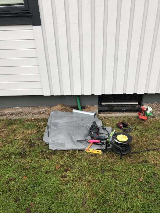 Grön kabelskyddsrör sticker upp bredvid husvägg med verktyg och byggmaterial på gräsmattan.