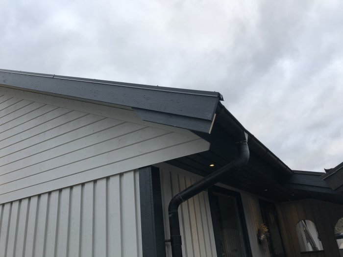 Husfasad med vit vindskiva och svart stuprör under ett gråmålat takutspring.