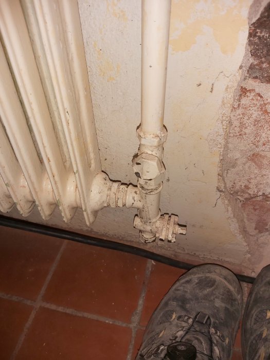 Radiator med synliga anslutningsrör och en stängd ventil, placerad intill en gammal vägg och klinkergolv.