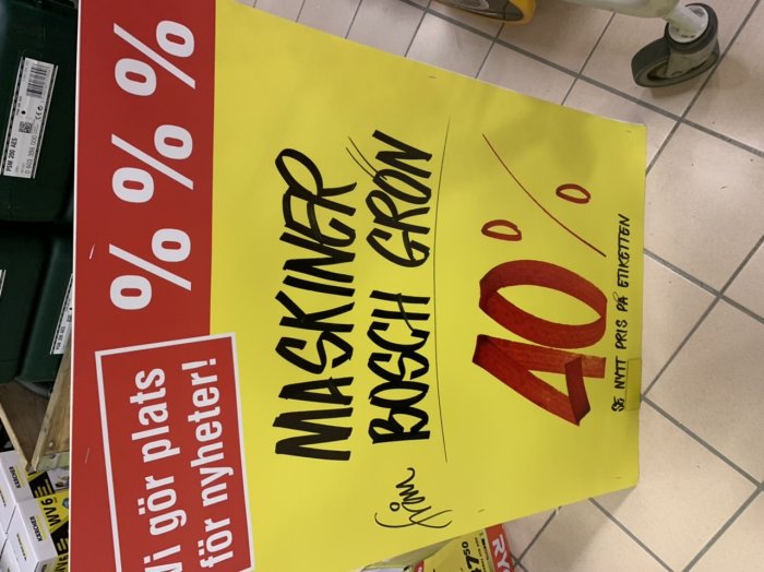 Gul skylt med text "MASKINER BOSCH GRÖN 40%" och en röd skylt med text "gör plats för nyheter!" på butiksgolv.