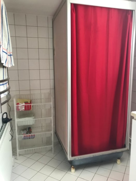 Äldre badrum med vitkaklade väggar och röd duschkabin, vit plasthylla och handduksvarmare.