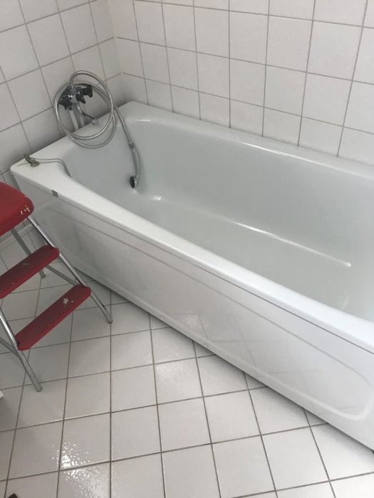 Äldre vitt badkar med matchande vita kakelväggar och golv i en badrumsinredning från 90-talet.
