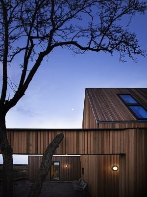 Modern träbeklädd byggnad vid skymning med tända utomhuslampor och en synlig måne på himlen.