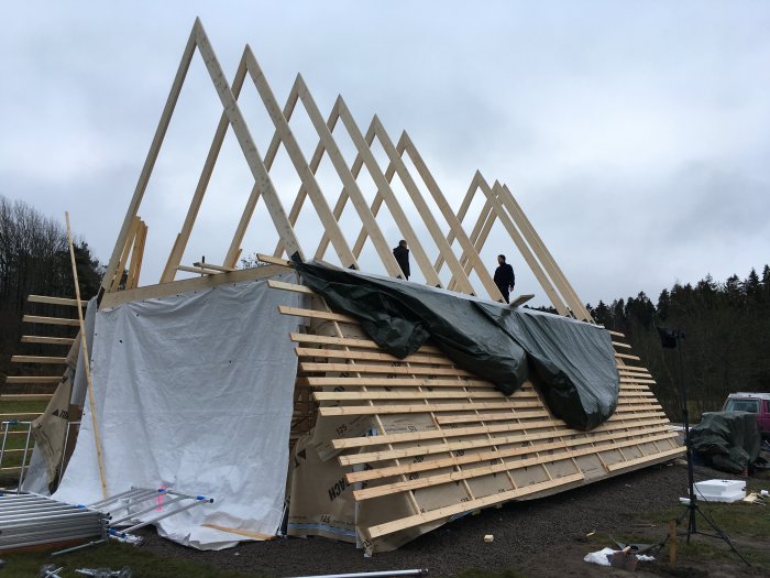 Takstolar för ett hus under uppbyggnad med personer som arbetar på konstruktionen, omgiven av skog.