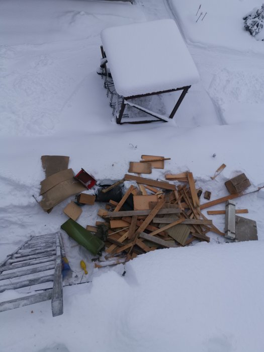 En hög med skräp och gammalt byggmaterial på snön, bredvid ett bord täckt av tjockt snötäcke.