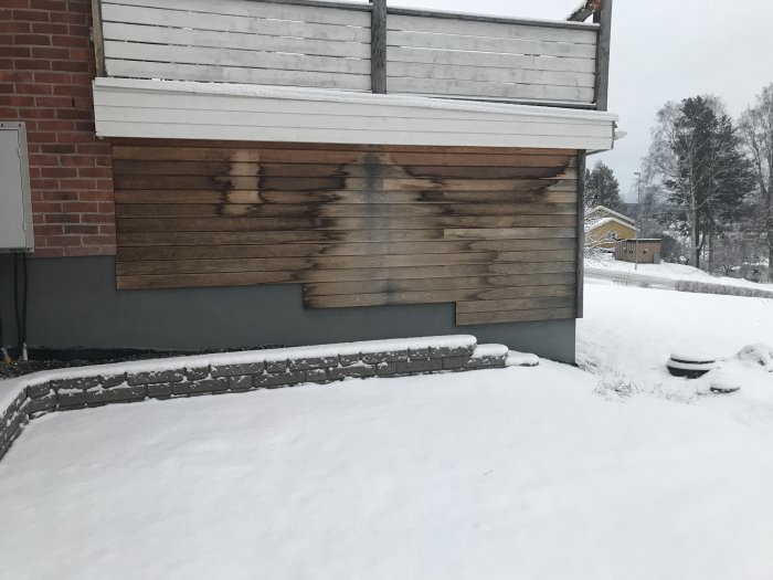 Delvis missfärgad träpanel på hus i snöigt landskap, variation i färg visar väderpåverkan.