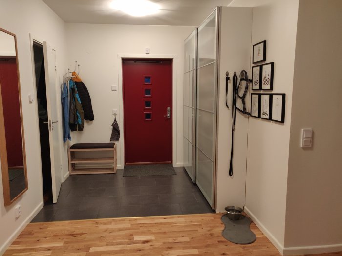 L-formad hall med vit vägg, röd dörr, stor garderob, träbänk, hängande jackor och ramar på väggen.