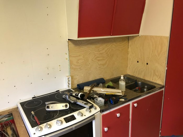 Kök under renovering med plywoodväggar, röda skåpluckor och verktyg på bänken.