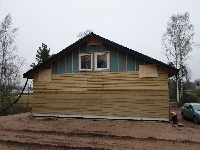 Nybyggt hus med pågående fasadarbete och träpanel, täckta fönster och lera runtomkring.