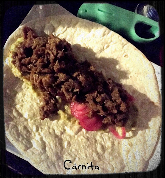 Hemlagad carnitas på tortilla med picklad rödlök och guacamole, serveringsklar med en grönsaksskalare i bakgrunden.