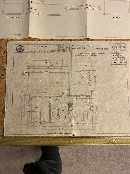 Fotografi av en arkitektritning för en husgrund, placerad på ett bord, med synliga måttangivelser och delar av en person.
