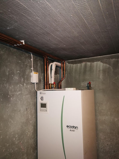 Ecodan värmepump i källare med kopparledningar och elektrisk utrustning på betongvägg.