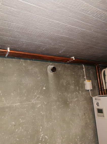 Värmerör löper längs betongvägg under träfärgat tak nära en luft/vattenvärmepump.