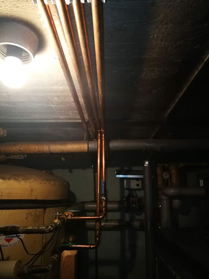 Rörinstallationer och en vattenpump inuti ett hus, med synliga kopparledningar och äldre rörsystem.