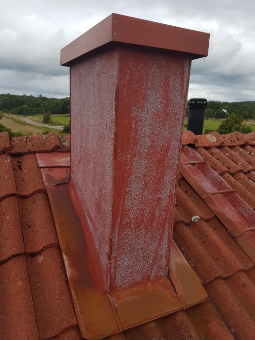Rostskyddsmålad ventilationstrumma på tak med kondens på utsidan, synlig mot bakgrund av tegeltak och landskap.
