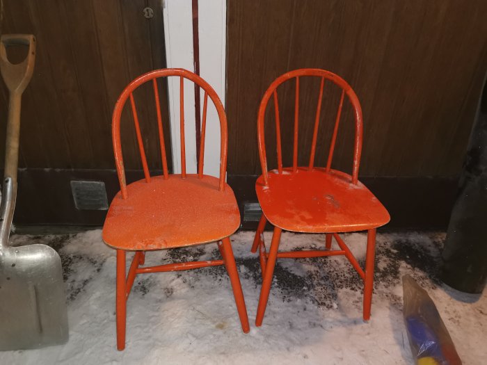 Två orange, slitet utseende köksstolar på en snöig bakgrund intill en spade och dagstidning.