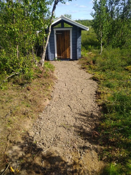 Nyanlagd grusgång som leder till ett grått uthus omgivet av gröna buskar och träd i sommarlandskap.