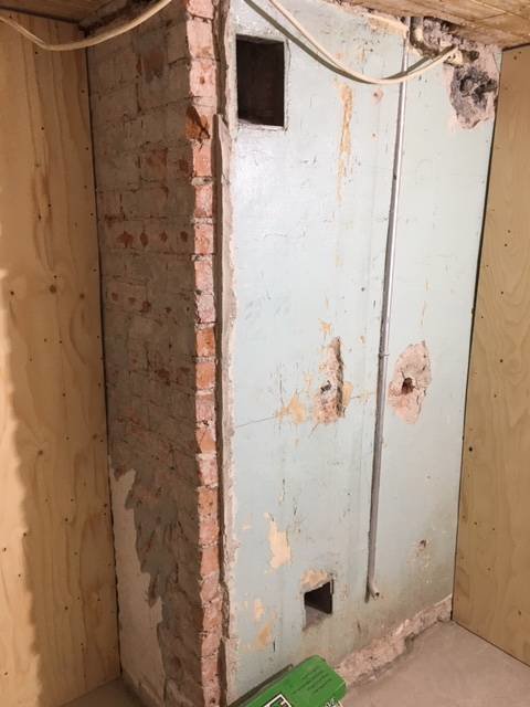 Hörn av ett pågående badrumsprojekt med oslipad murstock och väggar klädda med plywood och gips.