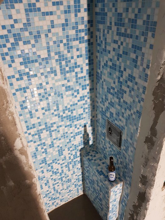 Renoveringsprojekt med blå och vit mosaikklinker på vägg och en ölflaska på en liten plattform.
