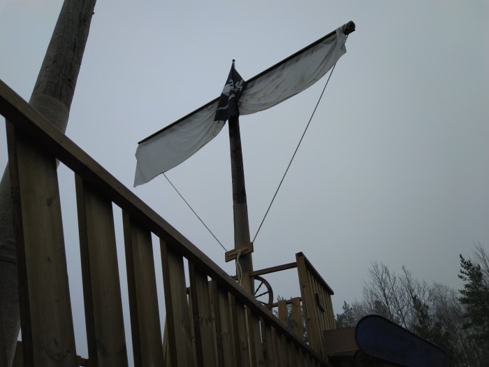 Nymonterat segel på mast vid trädäck, vitt segel mot grå himmel, omgiven av träräcken.