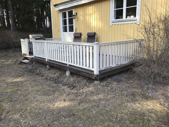 Gammal vitmålad trädäck och staket i dålig skick vid gul husvägg.