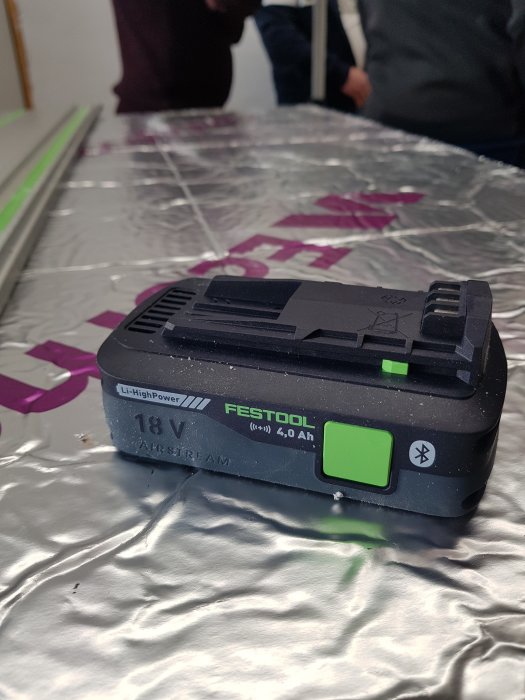 Ett kompakt 18V Festool litiumjonbatteri med 4.0 Ah kapacitet på en reflekterande yta, omgivet av oidentifierade personer.