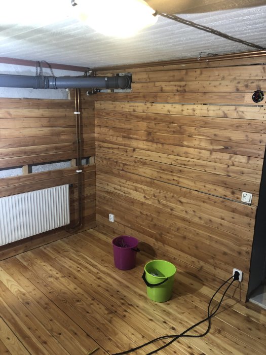 Källarrum under renovering med träpanelväggar, rör i taket och två hinkar på trägolvet.