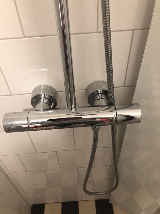 Kromad termostatblandare med anslutningskopplingar i dusch, vit kaklad vägg i bakgrunden.