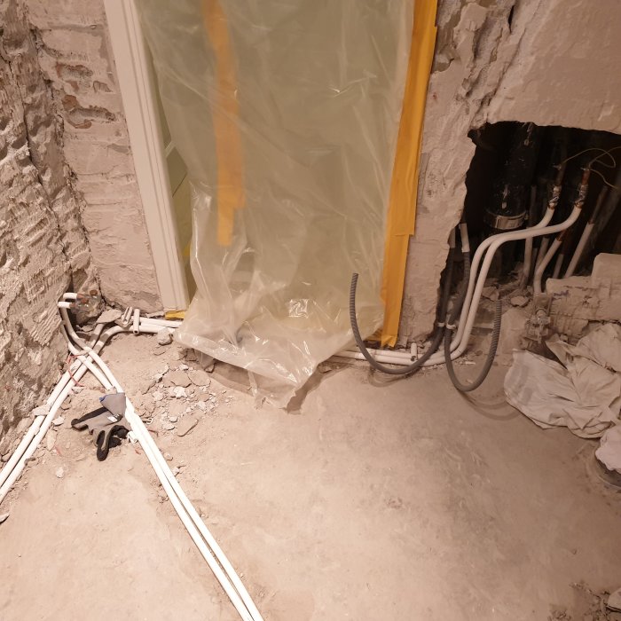 Rörmokeriarbete med synliga värmerör och kopplingar i ett under renoveringvänt rum med oslaget betonggolv och skyddsplast.