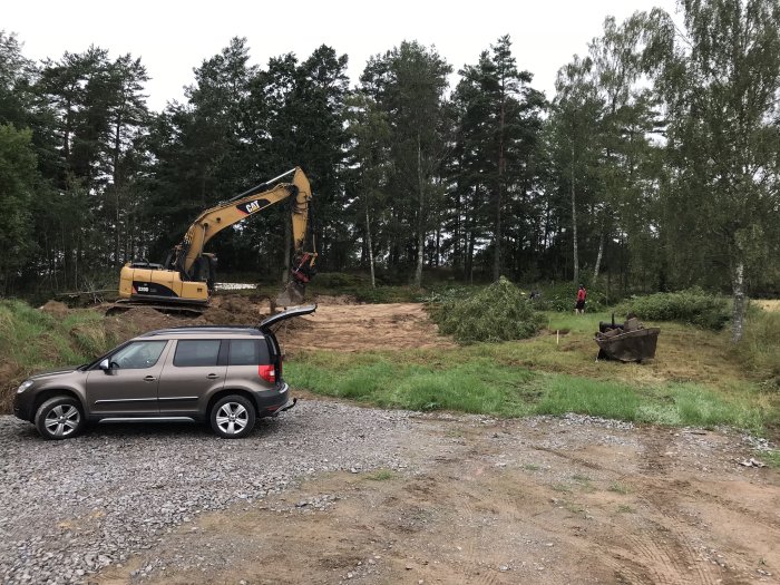 Grävmaskin schaktar mark för husgrund med skogsbakgrund, SUV parkerad i förgrunden.