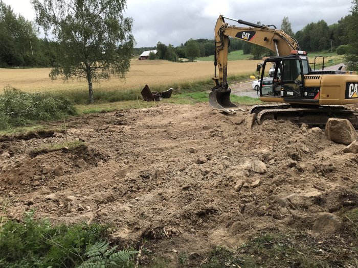 Grävmaskin på arbetsplats utför markarbeten för husgrund, med uppgrävt område och stenar synliga.