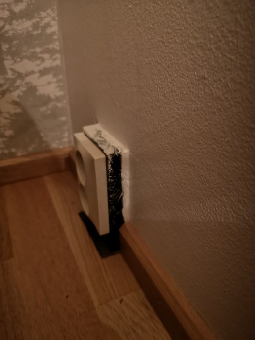 Delvis målat eluttag i brunt mot en texturerad vit vägg vid ett hörn ovanför ett laminatgolv.