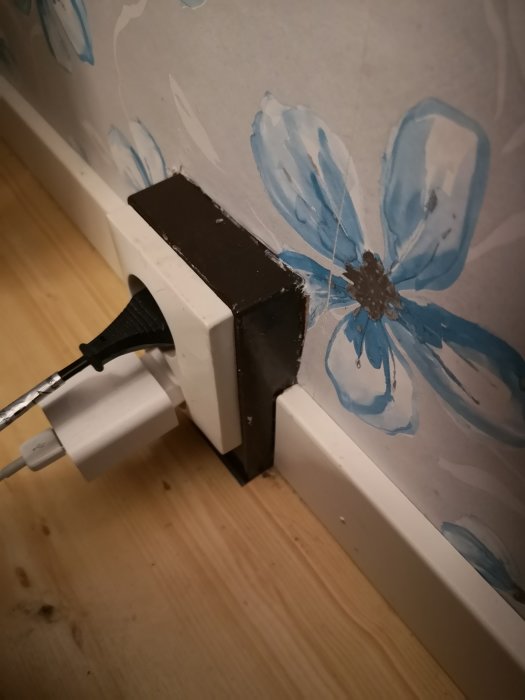 Omålat yttre av eluttag monterat på vägg med blommigt mönster, sladd inkopplad, nära en vit list.