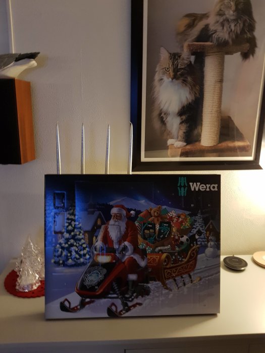 En adventskalender med julmotiv framför en inramad bild på en katt och en juldekoration på ett bord.