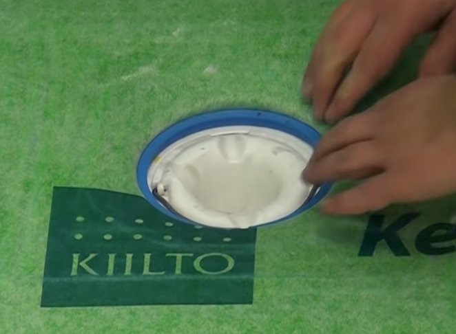 Hand som visar en grå klämring i en brunnsinstallation med Kiilto-reklam ovanför en grön bakgrund.
