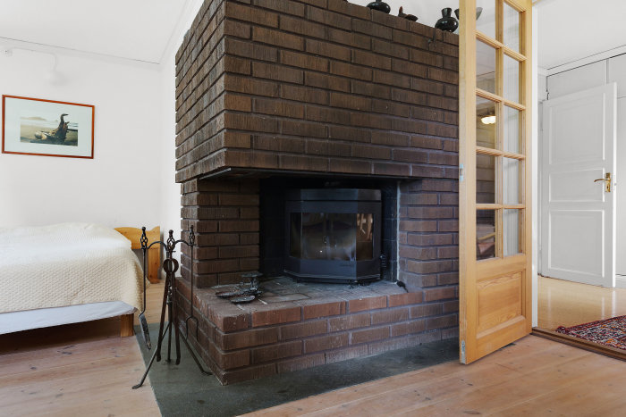 Murad eldstad med brun tegel och installerad kassett i ett hörn av ett rum med trädörr och vit vägg.