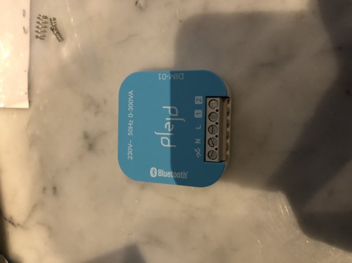 Blå Plejd-dimmerpuck upp-och-nervänd med anslutningsterminaler på vit marmorbakgrund.