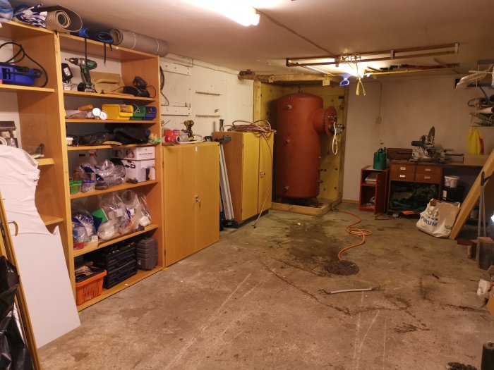 Oorganiserad källarlokal under renovering med öppna skåp och en stor röd tank.