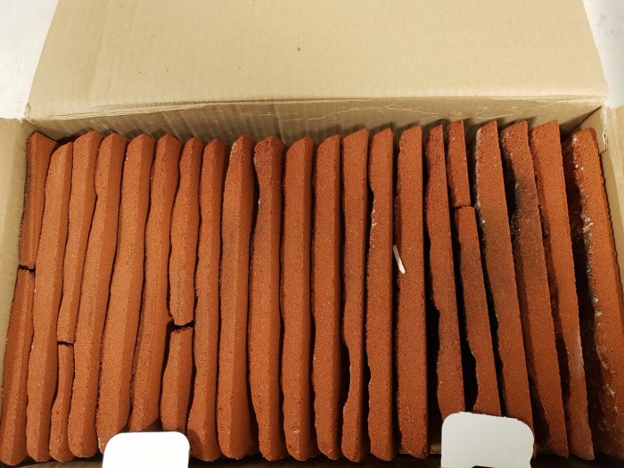 Öppen kartonglåda fylld med ordnade rader av rödbruna "fusktegelstenar", somliga med synliga sprickor.