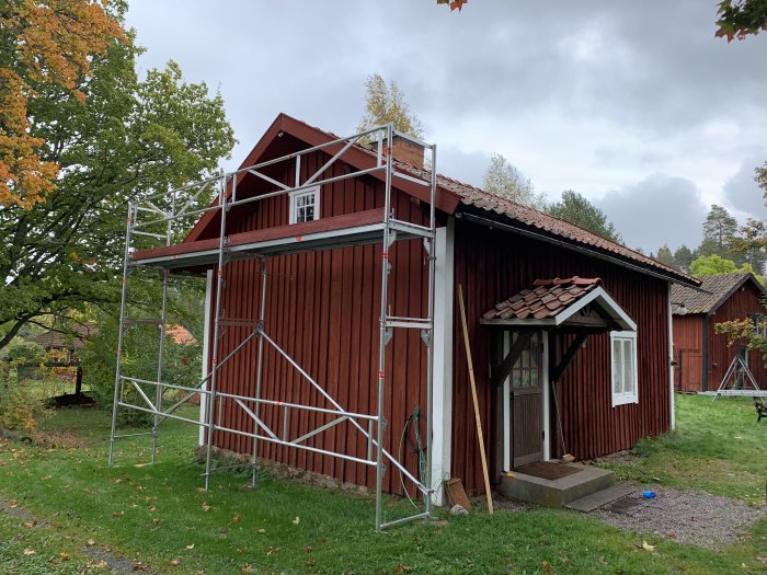 Renoverat rött trätorp med nytt tak och byggnadsställningar runt fasaden på en höstdag.