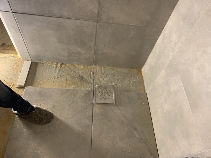 Delvis upptaget badrumsgolv med lösa plattor och synligt tätskikt vid en avloppsränna, fot i bild.
