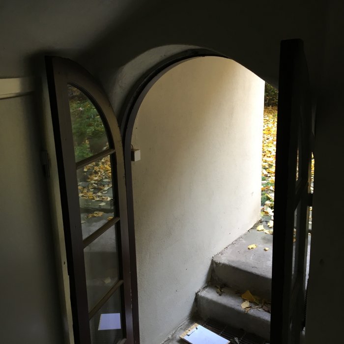 Ett trångt trapphus med en bågformad gårdsdörr som öppnar mot en innergård med höstlöv på marken.